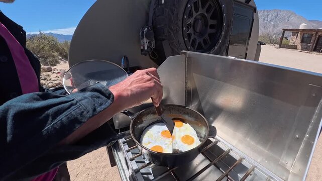 Mature man cooks eggs on a stove at a beach camp in Baja California using a teardrop trailer for travel and convenience.