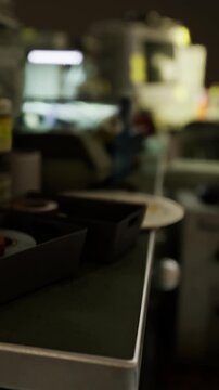 In a busy workshop, tools and materials are scattered on a table. The workspace shows signs of ongoing projects under artificial lighting. The atmosphere is active and engaged.
