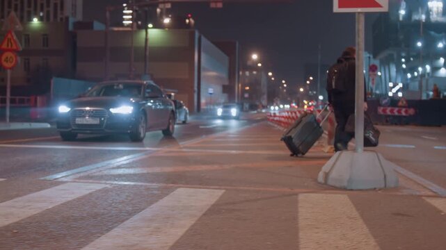 Night crosswalk with suitcase and headlights, lone traveler drags rolling luggage across striped pavement while cars wait with bright beams, construction cones and building lights frame scene,