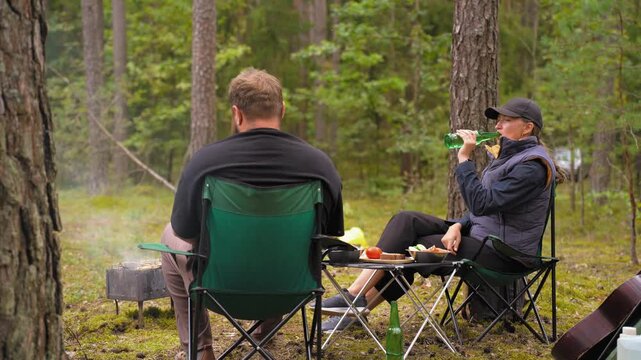 Man and woman relaxing in camping chairs, enjoying beer and snacks while having a pleasant conversation during a picnic in a beautiful pine forest, with smoke rising from a nearby barbecue