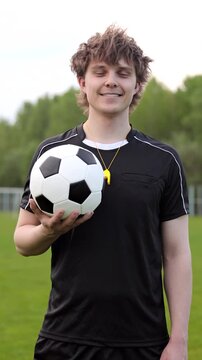 Young smiling soccer referee or coach holding a football in black referee uniform. Concept of fair play, sports rules and game control. Vertical video.