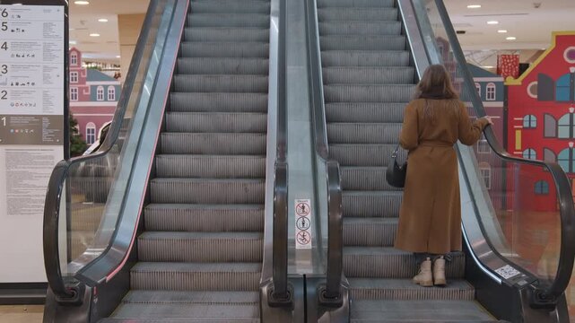 Shopper ascending mall escalator, brown coat. Handbag at side, slow deliberate steps toward upper level, directional signage and colorful storefront mural in background, empty center corridor, soft