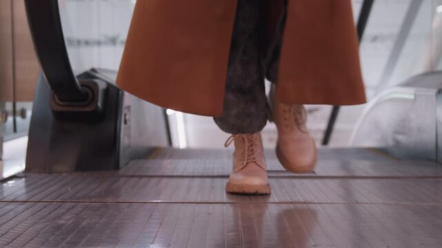 Caucasian woman brown boots on escalator, closeup of descending step, long coat hem swaying, textured metal tread, mall signage blurred, ambient warm lights, steady downward motion, commuter mood