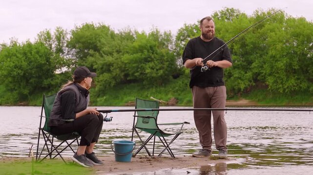 Mature man and woman enjoying a quiet day fishing together on the sandy bank of a river, sitting on folding chairs while patiently waiting for a catch on a peaceful, cloudy day