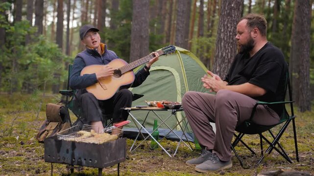 Happy young couple enjoying a romantic picnic in the forest with a barbecue, the woman playing an acoustic guitar while the man eats and listens to her song next to their tent