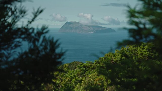 Cinematic Long Shot of Corvo Island on the Horizon Framed by Lush Green Foliage, viewed from Flores Island, Azores