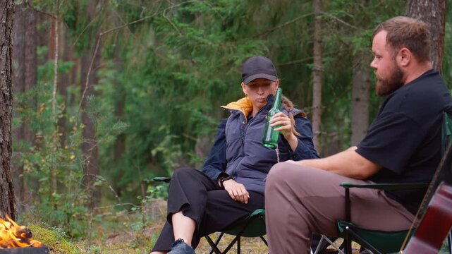 Young couple enjoying a quiet getaway, sitting on camp chairs, drinking beer from a bottle, and talking near a warm campfire in a tranquil pine forest during an autumn evening