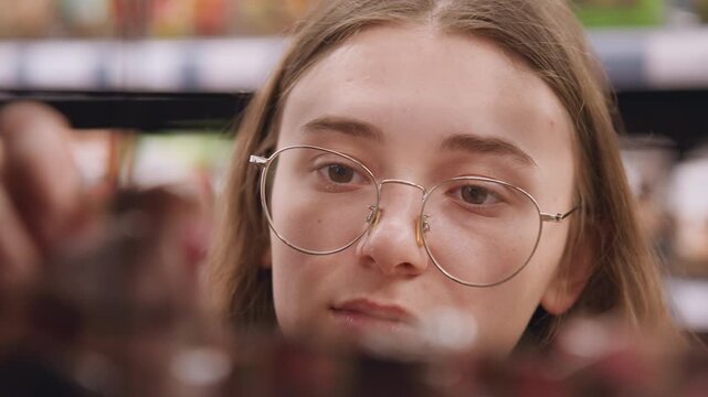 White woman comparing product choices on shelf with round glasses and steady gaze label reader checking nutrition, budget shopper weighing price, curious consumer reaching for snack jar while studying