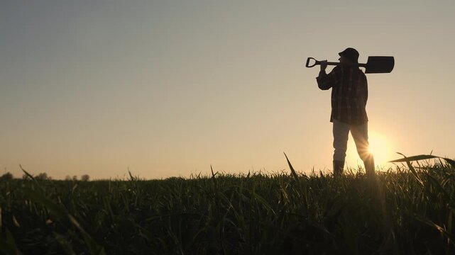 Silhouette of farmer standing in green agricultural field holding shovel at sunset during golden hour in countryside.