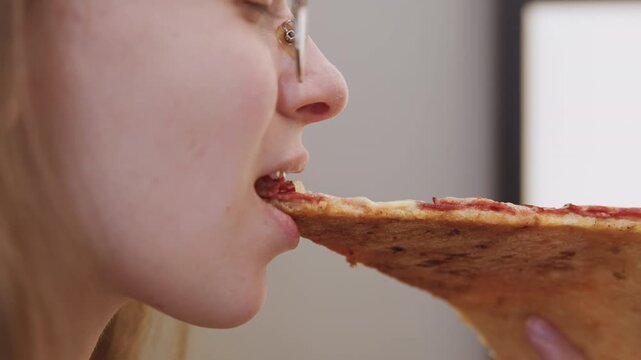 Caucasian woman bites pepperoni slice, glassesclad foodie tasting slice in studio setting, profile closeup on lips and mouth showing subtle smile and satisfied expression, warm indoor lighting