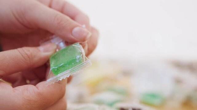 Closeup hands unwrapping green candy, transparent wrapper peeled to reveal sugar crystals, focused fingertips and careful peeling motion, blurred colorful candies in background, indoor countertop,