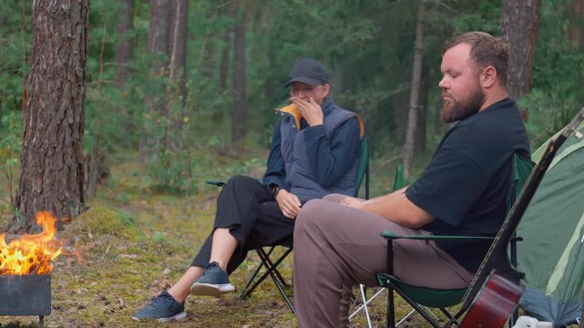 A young couple sits on camping chairs by the fire, drinking beer from green bottles and talking, and behind them in a beautiful pine forest setting there is a tent and a picnic table
