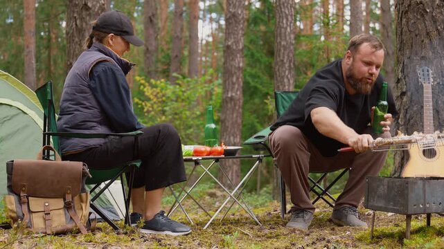 Happy young couple sitting on camping chairs in woods, talking and laughing together, grilling meat on barbecue picnic during their trip to the fresh air and drinking beer