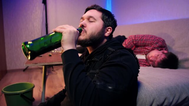 Side view of a bearded man drinking alcohol sits on the floor in living room, drinks alcohol from a bottle, feels nauseous and vomits into a green bucket. The party ended with alcohol intoxication