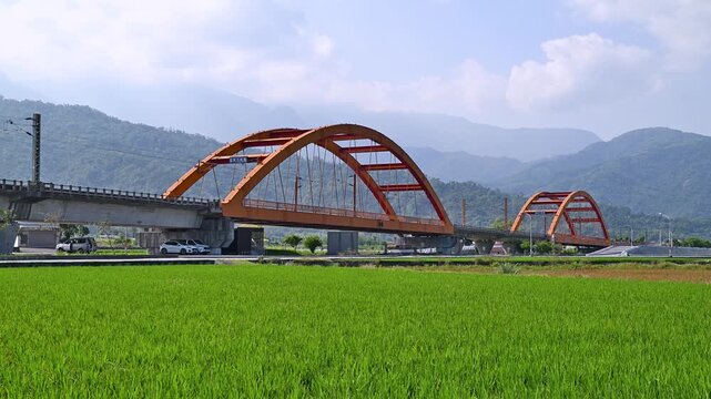 A high-speed train travels across a vibrant red arch bridge in Yuli, Hualien, Taiwan, set against a backdrop of lush green rice fields and mountains.