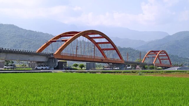 A passenger train with four silver carriages travels across a vibrant red arch bridge in the rural landscape of Yuli, Hualien, Taiwan.