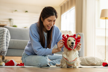 Happy beautiful young Asian woman dressing her pet dog in red reindeer costume sitting on living room floor smiling brightly enjoying playful holiday season preparation indoors © 220 Selfmade studio