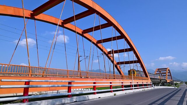 A silver train travels across a vibrant red steel arch bridge in Yuli, Hualien, Taiwan, set against a clear blue sky and mountain backdrop.