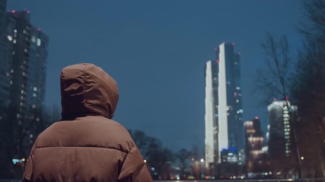 Hooded figure faces illuminated city skyline in brown puffer jacket at riverfront park, distant highrises reflecting in water, quiet cinematic mood with subtle bokeh lights, chilly evening pause