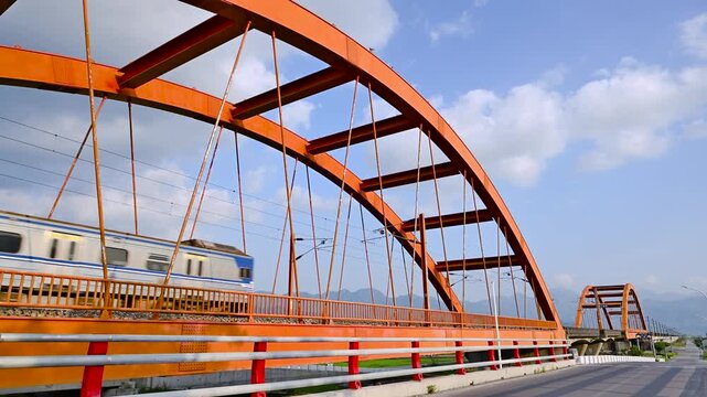 A passenger train with four silver carriages travels across a vibrant red steel arch bridge in Yuli, Hualien, Taiwan, under a clear blue sky.