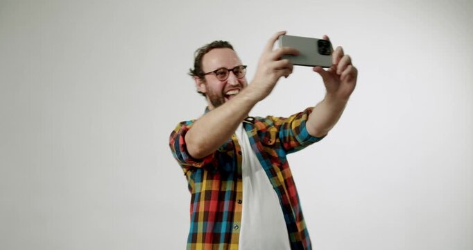 Happy man taking a selfie with his smartphone. He is smiling and laughing while posing for the camera. The man is wearing a colorful plaid shirt and glasses. He looks fashionable and casual.