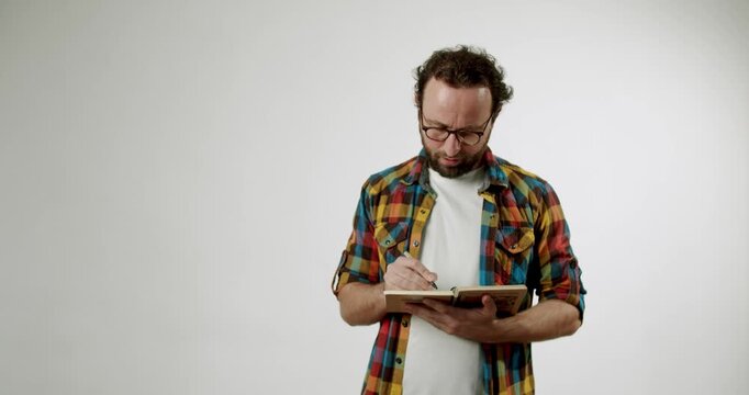 A man in a plaid shirt writes in his notebook. He wears glasses and has a beard. He stands against a white background. This video evokes feelings of creativity, thoughtfulness, and concentration.