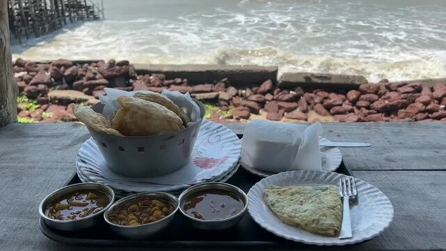 Indian breakfast with puri sabji, bread butter, and omelet served with sea waves in the background; professional travel and food photography of an oceanfront morning meal, beachside dining, and coasta