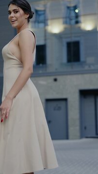 Stylish woman in a beige dress poses for the camera. She twirls, looks over her shoulder, smiles, kicks up her heel, and spins. Urban fashion shoot with modern architecture backdrop.