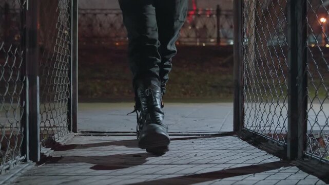 Closeup boots walking through gate, heavy leather boots step over threshold onto cracked pavement, metal chain link sways, shadowed sidewalk lit by distant streetlight, rhythmic footsteps echo