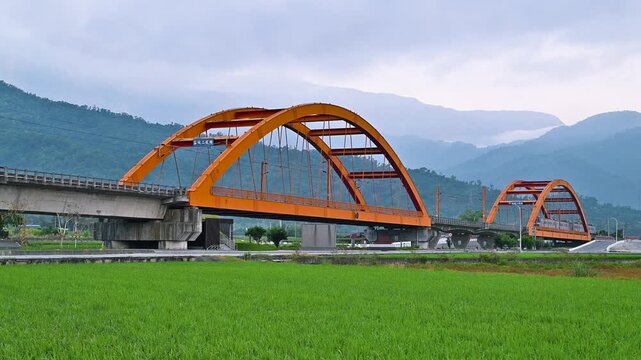 A passenger train with four silver carriages travels across the iconic orange arched Ke-Cheng Bridge in Yuli, Hualien, Taiwan, surrounded by lush green rice fields and mountains.