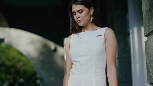 A young woman in a stylish white dress poses near a building. She exudes confidence and elegance, capturing a sense of urban chic. Golden earrings add a touch of glamour to her look.