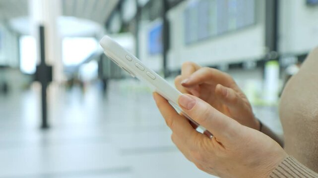 Close-up of hands holding smartphone in public space. Background shows blurred indoor area with pillars and screens. Person wears light-colored sweater. Scene suggests waiting or traveling