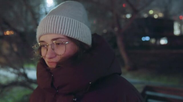 White woman thoughtful smile under lights in park, round glasses catching lamplight, knitted hat and puffer jacket, subtle expression changes from shy to playful, cinematic grain and soft nighttime