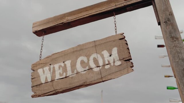 A rustic wooden welcome sign hangs from a weathered post, gently swaying in the breeze. White-painted letters pop against the aged wood. Glass bottles, repurposed as decorations, hang nearby.