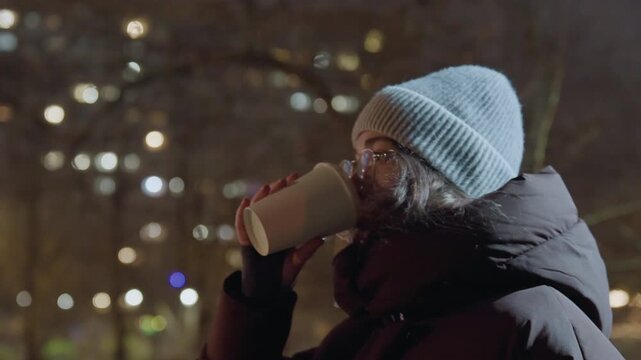 Night park woman sipping coffee bundled in puffer coat, wearing beanie and glasses under soft streetlight bokeh, contemplative gaze toward skyline, slow breath visible in cold air, hands warming cup,