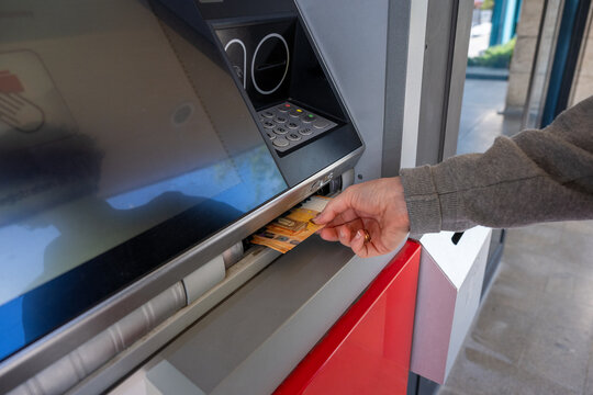 A person's hand reaches out to take a bundle of Euro banknotes from the cash dispenser slot of an automated teller machine (ATM).