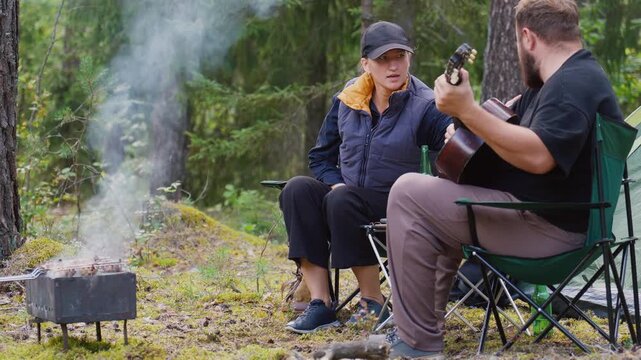 Young couple enjoying a picnic in the woods, with a bearded man playing an acoustic guitar while the woman sings along near a bonfire, a tent, and a small table with fresh vegetables