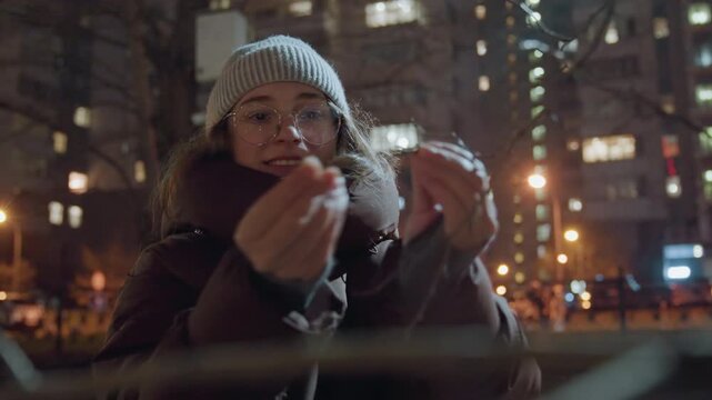 Woman smiling as she fixes ribbon outdoors. Charming woman in winter evening setting work indoors. Woman happily tying ribbon on fence during winter evening with glowing streetlights