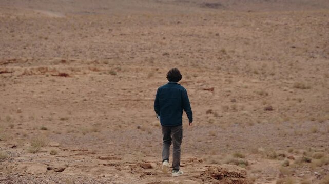 A young man walks through a vast desert landscape in Morocco. The warm colors of the sand and rocks contrast with his dark clothing as he explores the rugged terrain.