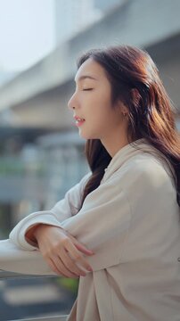 Happy Asian Gen Z woman smiling outdoors in the city. Beautiful young female with long hair enjoying a sunny day, showcasing wellness, positive emotions, and a modern lifestyle.