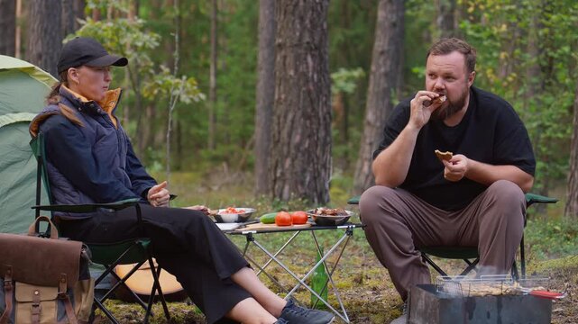 Young man and woman relaxing in camping chairs by a tent in the woods, enjoying a picnic with beer and grilled food while sitting around a campfire in the serene natural environment