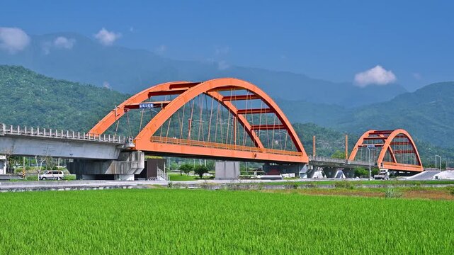 A vibrant orange arch bridge spans across a lush green rice field in Yuli, Hualien, Taiwan, with a train crossing under a clear blue sky.
