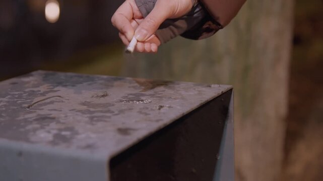 Gloved hand dropping cigarette into metal ashtray, closeup focus on fingers releasing lit stub, rusted wet surface, nighttime park setting, muted streetlight glow, quiet solitary mood, routine