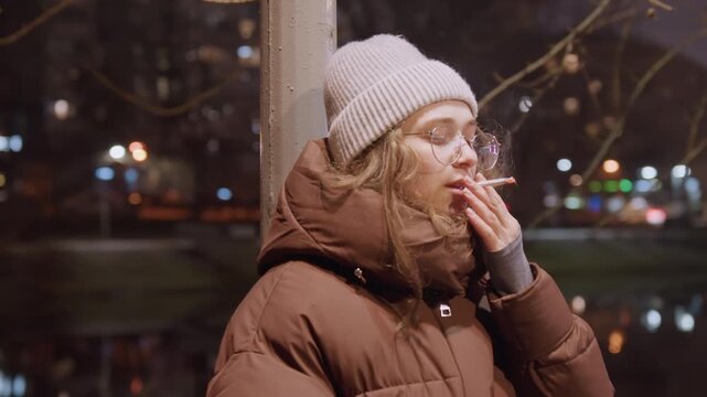 Young woman smoking by river. Latenight portrait of solitary figure in puffy coat and knit hat, glasses catching soft streetlight, cigarette lit, breath visible in cold air, hand to mouth, exhale