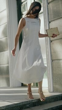 A young woman in a white dress and gold heels exits a building. She adjusts her shoe while holding a gold clutch. The sun shines, and her hair blows in the breeze.