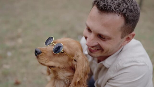 A man and his Cocker Spaniel enjoy a sunny day in the park. The dog wears round sunglasses, tilting its head. The owner smiles at his pet. This evokes feelings of happiness, companionship,