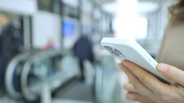 Person holds phone while standing near escalator. Background shows blurred commuters moving. Focus remains on phone in hand. Indoor public space with bright lighting. No animals or plants visible
