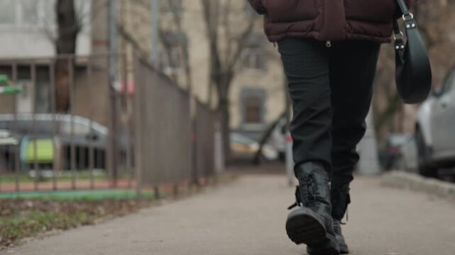 Closeup of caucasian woman walking along city sidewalk wearing black boots with laces and carrying small purse, muted winter tones, steady stride on textured pavement near parked cars and metal fence,