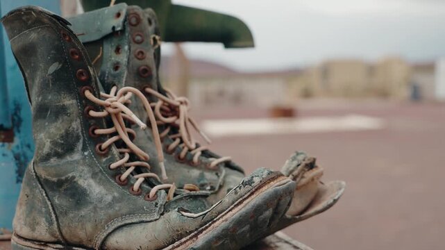 Close-up of a pair of worn-out, dusty work boots resting on a metal surface. The boots show significant wear and tear, with visible cracks in the leather and missing sections of the sole.