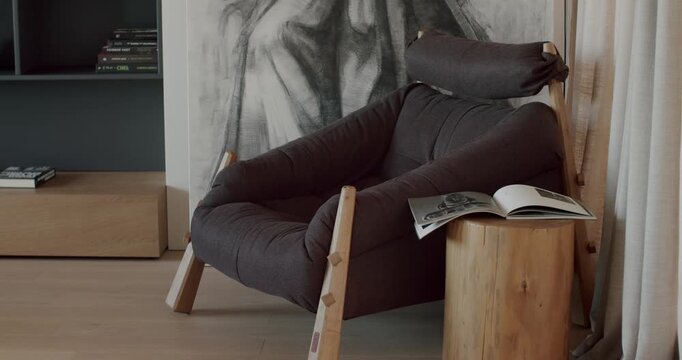 Comfortable modern armchair in a stylish living room. Charcoal-colored armchair with wooden legs, wood side table, a book and charcoal drawing. Calming, cozy, and comfortable setting.